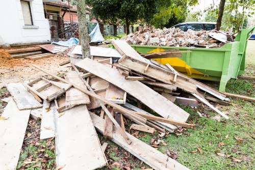 A pile of discarded wooden boards and planks, some with nails and paint remnants, lying on a grassy area beside a green skip filled with broken bricks and rubble. The boards are weathered, with rough textures, chipped edges, and varying shades of light brown and beige. The skip, positioned on a lawn, appears to be part of a rubbish removal operation, with part of a residential property and a tree visible in the background. This scene is typical of outdoor waste clearance in a suburban area, potentially in Colindale, with the clutter indicating renovation or construction debris that a company like Commercial Waste Removal Colindale may handle for commercial or residential clients.