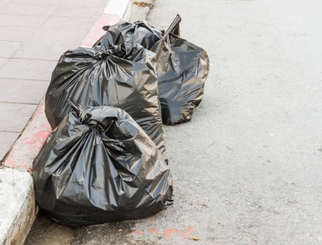 Two large black plastic rubbish bags filled with waste materials are placed on a pavement next to a curb in an outdoor urban environment. The bags appear to be tightly knotted at the top and are slightly bulging, indicating they are full. One bag is positioned closer to the foreground, partially resting on a concrete curb with visible red and white painted markings along the edge, while the other bag is situated slightly behind and to the right. The surface beneath the bags is a grey asphalt street, and the background shows a paved footpath with a slight texture. This scene is typical of a rubbish removal situation in a residential or commercial area, with the presence of waste collection activities likely handled by services such as Commercial Waste Removal Colindale, serving local areas in London postal districts.