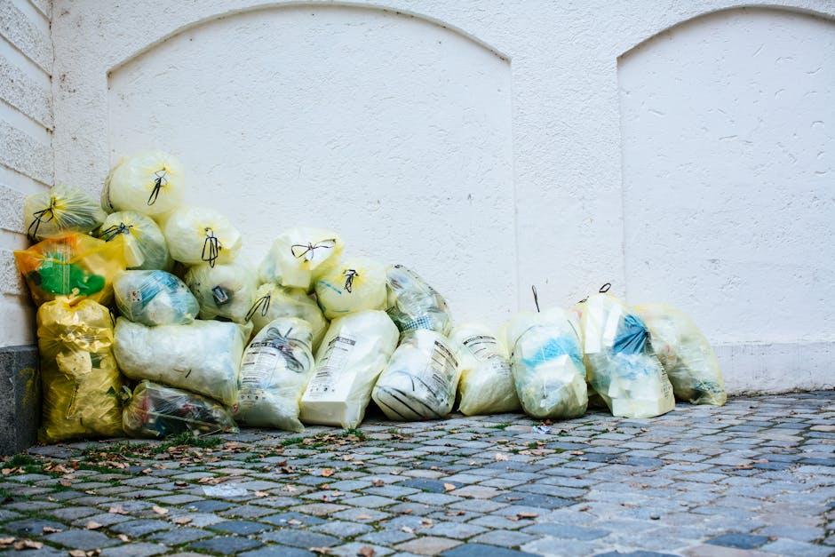 A person dressed in a plaid shirt, green gloves, and grey trousers is holding open a large black rubbish bag made of shiny plastic material, positioned outdoors on a grassy area. The individual is sta
