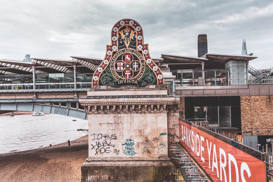 A large decorative coat of arms mounted on a stone pillar of a bridge structure over the River Thames in London. The crest features a detailed emblem with various symbols, including a crown at the top