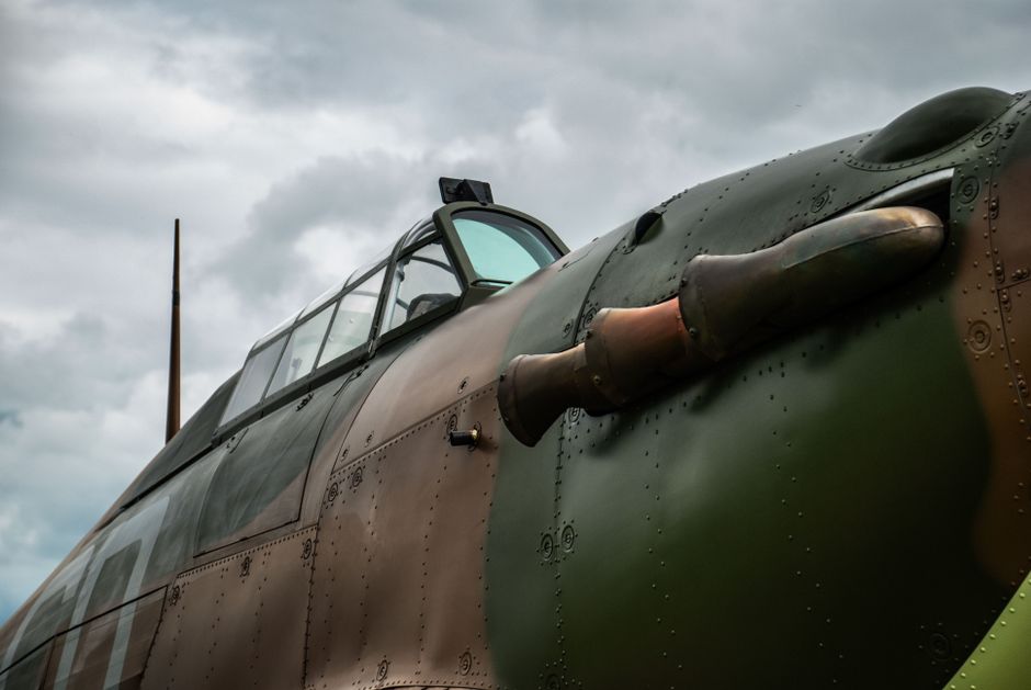 Close-up image of a vintage model aircraft, displaying the cockpit and upper fuselage, which is painted in a faded green camouflage pattern with signs of wear and weathering. The aircraft features a c